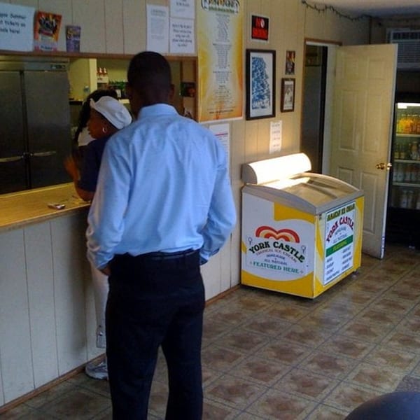 A man in a blue shirt stands in a Jamaican restaurant with a woman behind the counter.