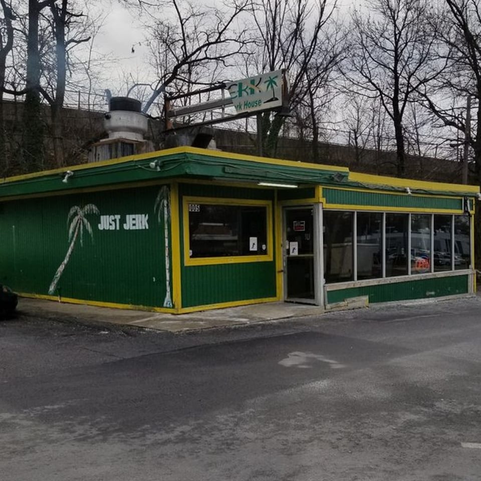 Just Jerk restaurant exterior, a green building with yellow trim and palm tree murals, under a cloudy sky.